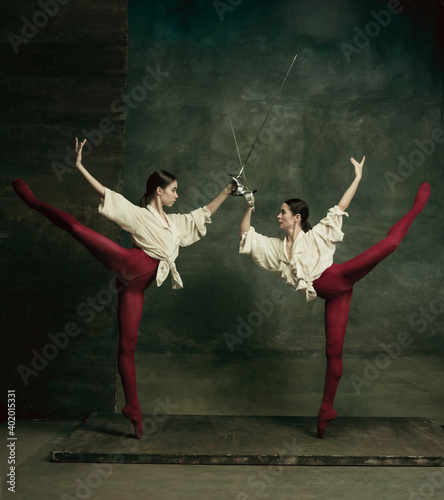 Flying. Two young female ballet dancers like duelists with swords on dark green background. Caucasian models dancing together. Ballet and contemporary choreography concept. Creative art photo. photo