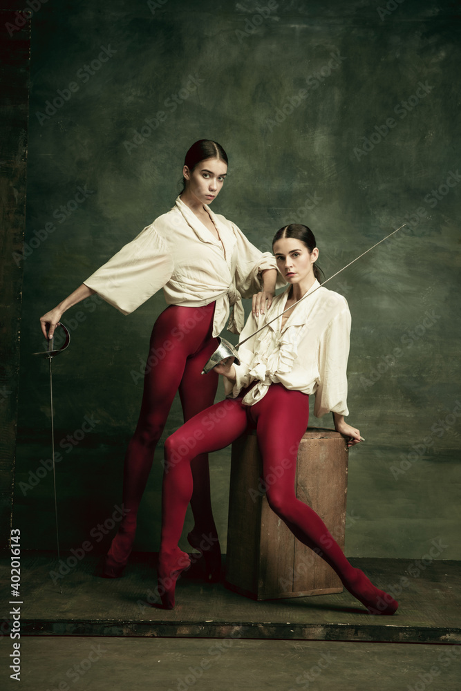 Enemies. Two young female ballet dancers like duelists with swords on dark green background. Caucasian models dancing together. Ballet and contemporary choreography concept. Creative art photo.