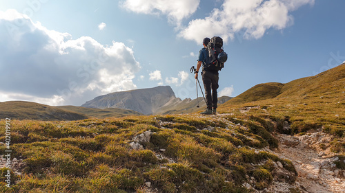 A man with a big hiking backpack hiking on a plateau in high Italian Dolomites. There are golden mountain slopes around the pathway. High and sharp mountains around. Remote and raw area. Calmness photo