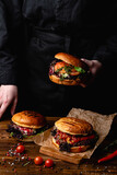Chef in a black apron holding a juicy burger, with a table standing in front of him, hands only visible