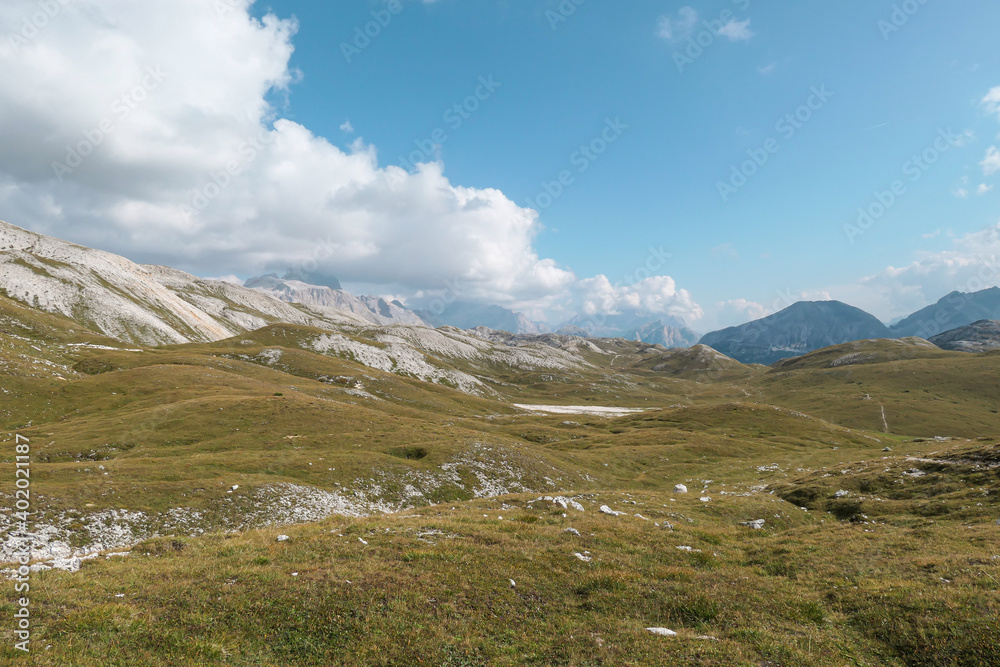 A panoramic view on a high plateau in Italian Dolomites. There are high mountains around. The area is partially overgrown with grass and partially covered with stones and pebbles. Remote and desolate
