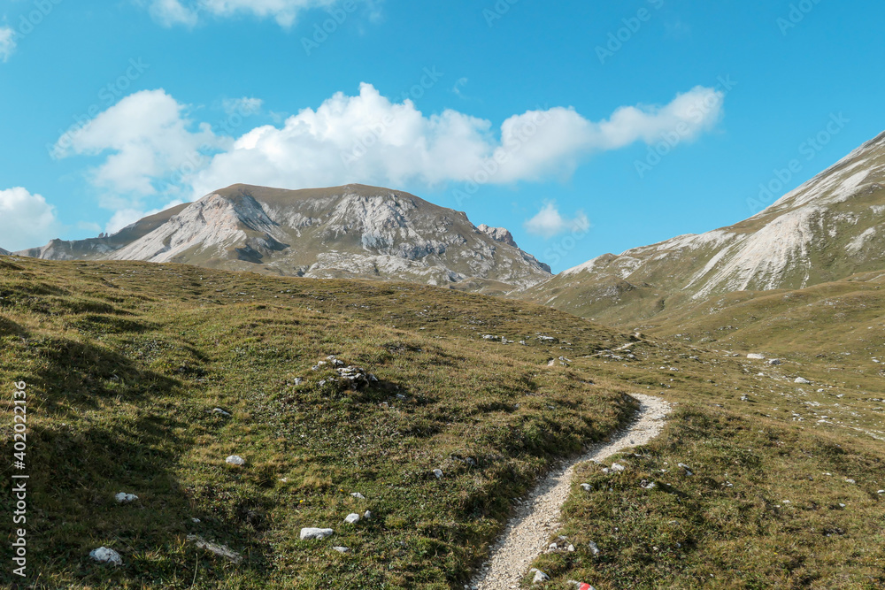A narrow pathway along a high plateau in Italian Dolomites. Stony landscape, with a bit of green grass. High mountain chains in the back. Desolated and remote landscape. Natural habitat. A few clouds