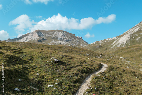 A narrow pathway along a high plateau in Italian Dolomites. Stony landscape, with a bit of green grass. High mountain chains in the back. Desolated and remote landscape. Natural habitat. A few clouds