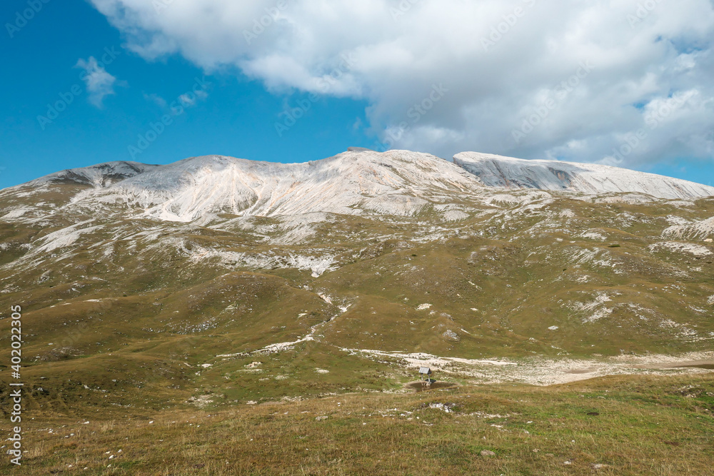 A panoramic view on a Seekofel peak, towering above a high plateau in Italian Dolomites. The area is partially overgrown with grass and partially covered with stones and pebbles. Remote and desolate