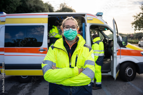 Portrait of young millennial woman with arms crossed at first aid in front his colleagues in ambulance at break time at pandemic time from Coronavirus, Covid-19 He wearing face mask and uniform work