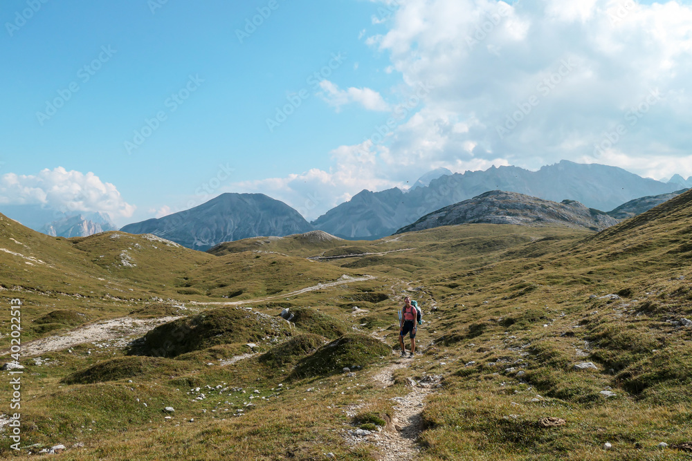Fototapeta premium A woman hiking along a high plateau in Italian Dolomites. High mountains around. The area is partially overgrown with grass and partially covered with stones. Remote and desolate area. Freedom