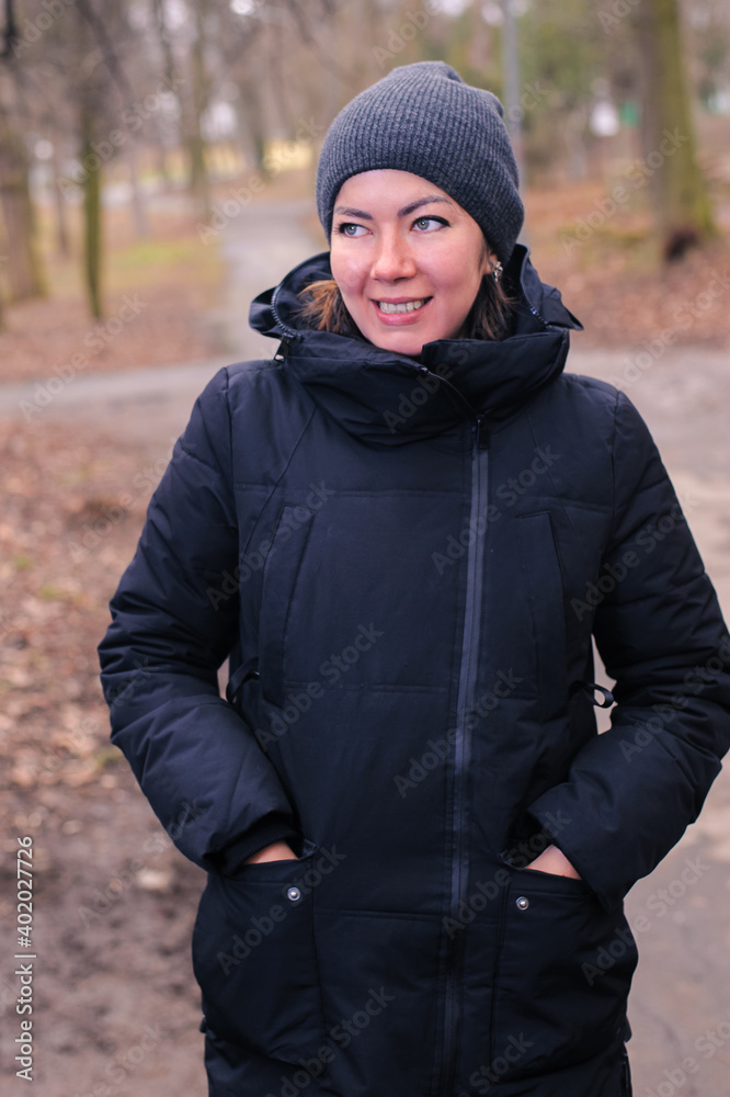 Smiling beautiful young woman in warm winter black clothes and knit cap standing in the park Smiling beautiful young woman in warm winter black clothes and knit cap standing in the park