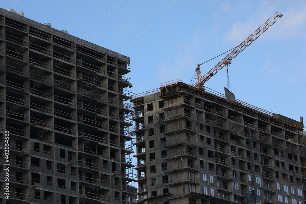 Two new concrete multi-storey buildings under construction with a crane lifting the construction plate.Gray high-rise structure with windows and ceilings in the working stage against the blue sky