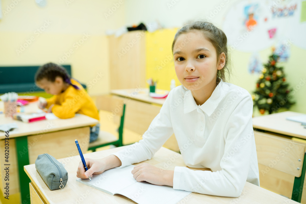 Little girl in a white shirt with gathered hair sits at a desk and writes in a notebook Little girl in a white shirt with gathered hair sits at a desk and writes in a notebook