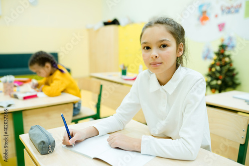 Little girl in a white shirt with gathered hair sits at a desk and writes in a notebook