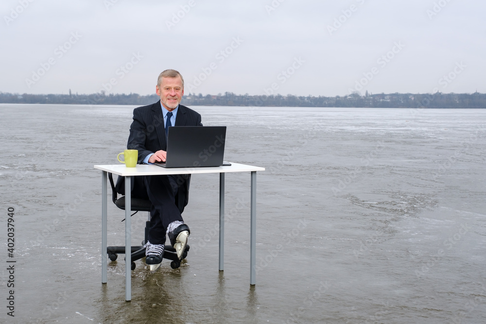 elderly bearded businessman in suit and skates works with laptop on table and sitting in chair in the middle of a frozen lake. Copy space for text, work on vacation concept