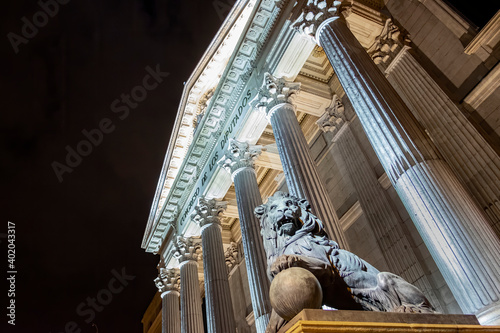 night view of the main facade of the Palacio de la Cortes, seat of the Congress of Deputies in Madrid, Spain, with the columns and one of its iconic bronze lions in the foreground
