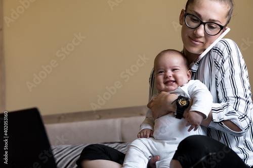 Childhood, motherhood, family concepts - successful smiling rich business woman Lady mother sit on bed with infant child baby talking on phone working on lap top computer at home in distance indoors