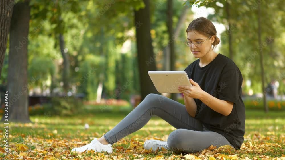 Slow Motion Young woman in glasses sits on the lawn in autumn city park and works on tablet computer Slow Motion Young woman in glasses sits on the lawn in autumn city park and works on tablet computer