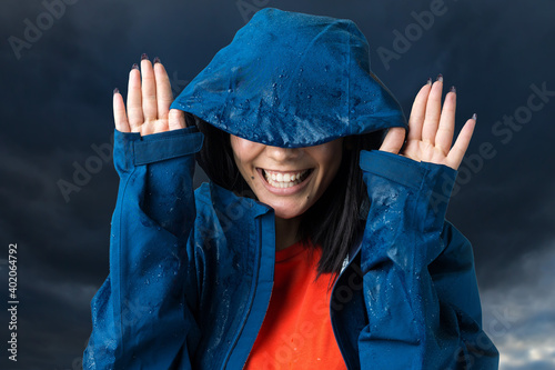 Portrait of a smiling girl dressed in blue raincoat in drops posing with hood against the background of the dramatic sky. Bad weather concept. photo