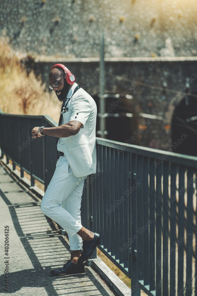 A vertical shot of a fashionable bearded bald black guy in a white costume waiting for his friend and looking at his watch, while leaning against iron fencing of a pedestrian bridge over the railroad A vertical shot of a fashionable bearded bald black guy in a white costume waiting for his friend and looking at his watch, while leaning against iron fencing of a pedestrian bridge over the railroad