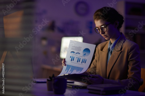 Young female finance manager sitting at desk looking through papers late at night in office room, copy space