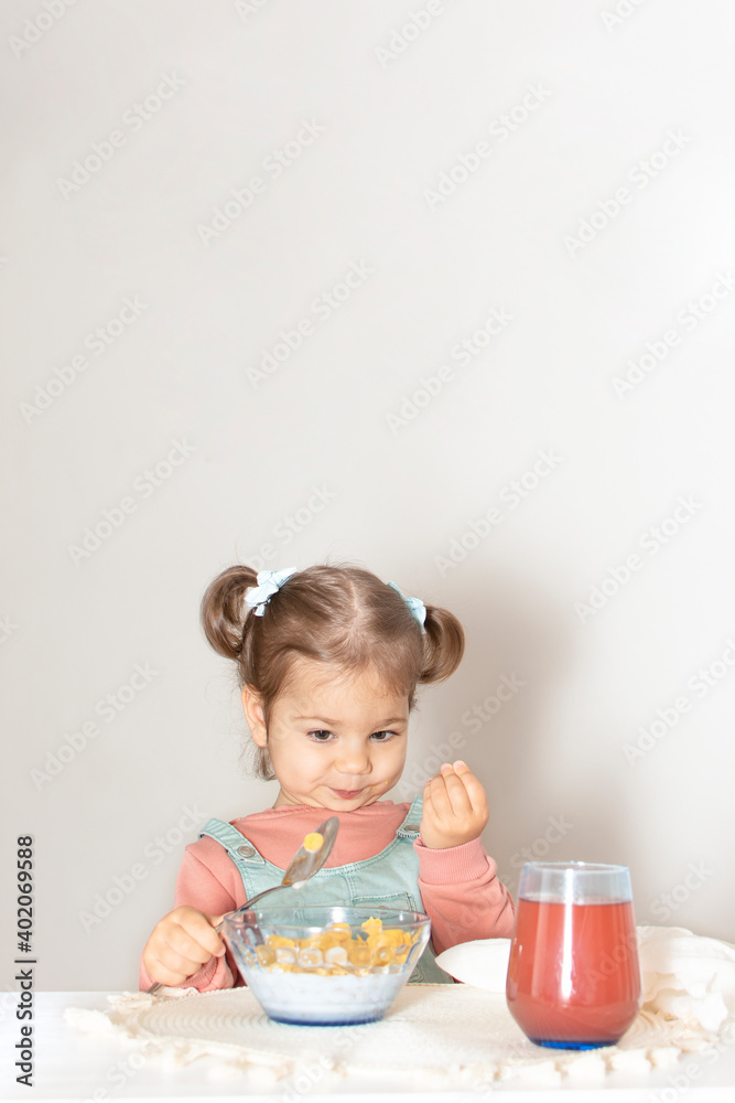 Adorable cute baby girl eating corn flakes for breakfast. Beautiful kid eat cereal with the milk in the kitchen. Healthy nutrition for small children. White background and copy space for text. Adorable cute baby girl eating corn flakes for breakfast. Beautiful kid eat cereal with the milk in the kitchen. Healthy nutrition for small children. White background and copy space for text.