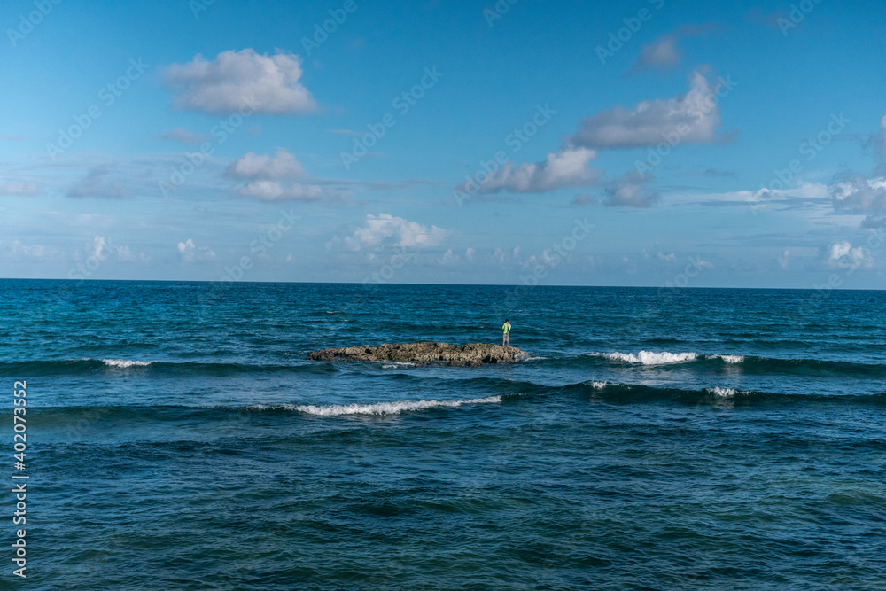 man fishing on a small island in the middle of the ocean with blue sky and some clouds