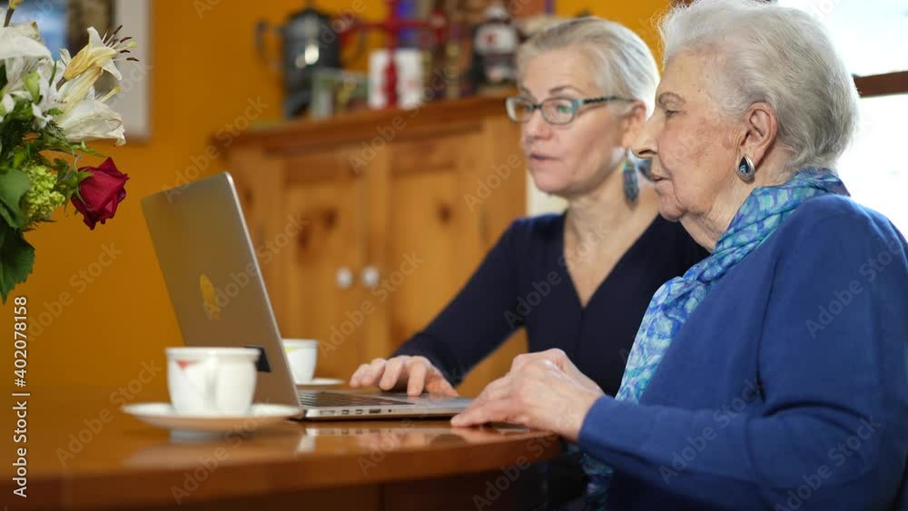 Low angle view of mature woman and elderly mother working on laptop computer on dining room table having tea.