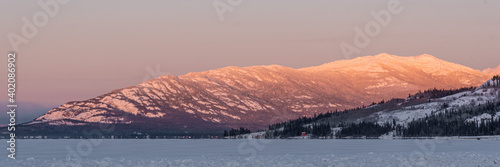 The sun rising over a huge snow capped mountain on a crisp winter morning with clear  bright sky  orange mountains peak with frozen lake and woods in foreground. 