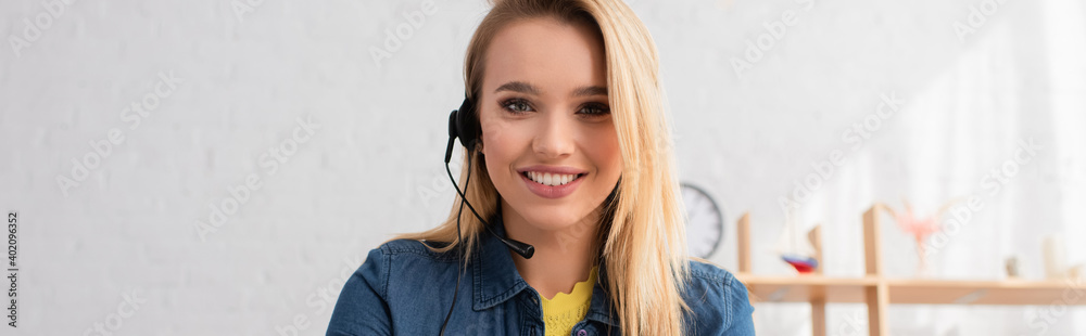 Portrait of blonde woman in headset looking at camera at home on blurred background, banner Portrait of blonde woman in headset looking at camera at home on blurred background, banner