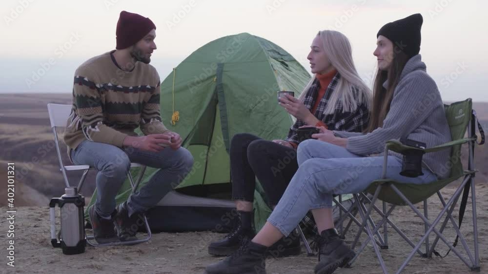 Young happy friends campers sitting near the tent and the car, drinks a hot tea and talks Young happy friends campers sitting near the tent and the car, drinks a hot tea and talks
