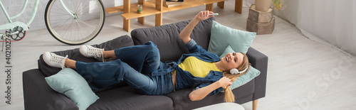 High angle view of blonde woman with smartphone laughing while lying on couch at home, banner