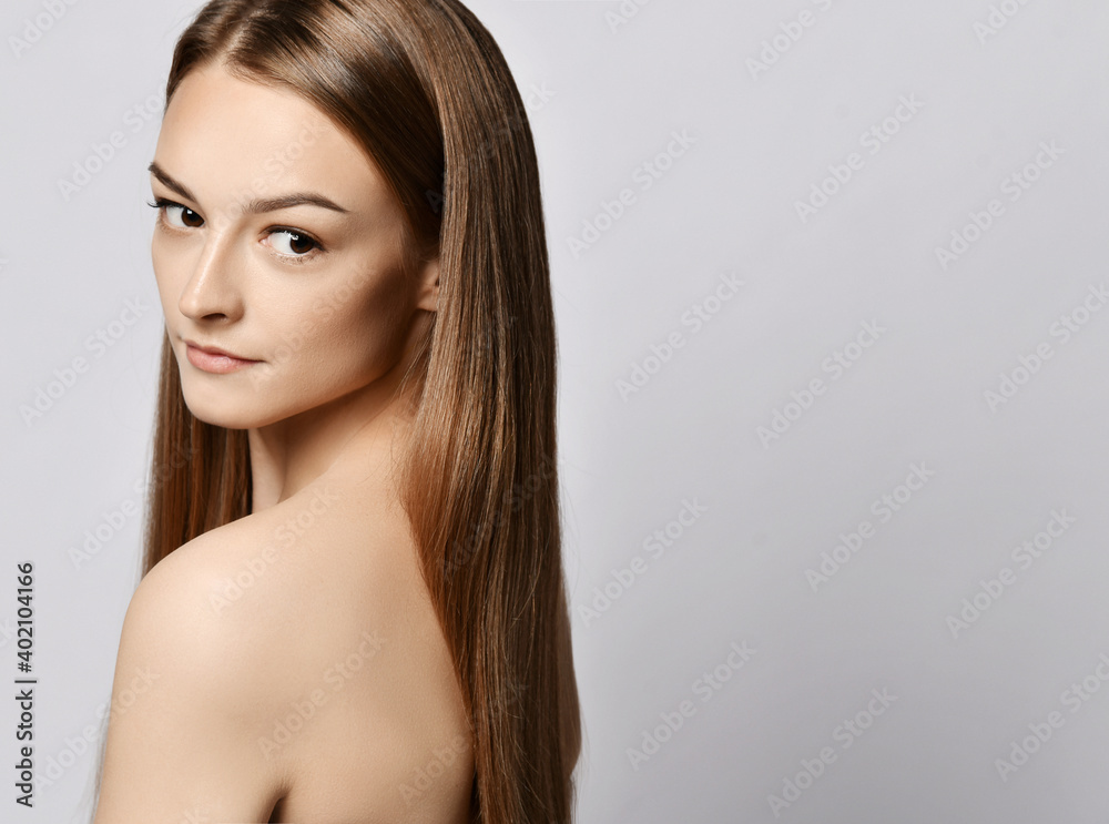 Portrait of sensual young slim woman with long silky straight hair standing with her back to camera looking over her naked shoulder at copy space over grey background. Haircare, beauty, wellness