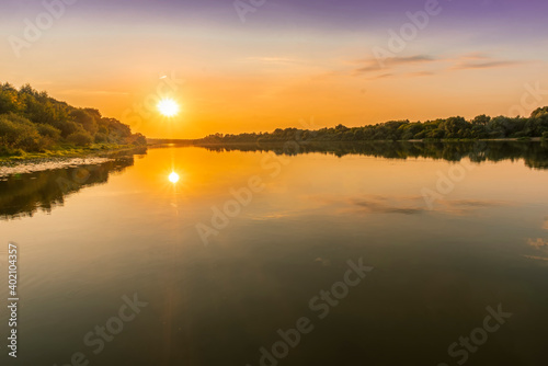 Fototapeta Naklejka Na Ścianę i Meble -  Scenic view at beautiful summer river sunset with reflection on water with trees , golden sun rays, calm water ,deep blue cloudy sky and glow on a background, spring evening landscape