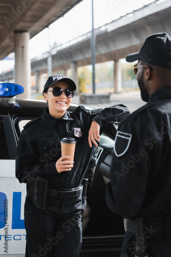 happy policewoman with paper cup looking at african american colleague while leaning on patrol car on blurred background.