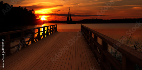 Sunset on the landing stage in Sweden, with the silhouette of a passing sailboat © Stefan