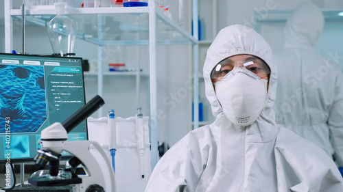 Microbiologist sitting in laboratory wearing ppe suit looking at camera in modern equipped lab. Team of scientists examining virus evolution using high tech and chemistry tools for scientific research