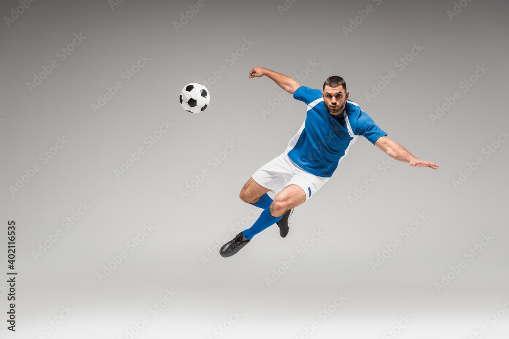 Fototapeta premium Sportsman looking at camera while jumping near football on grey background