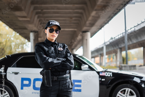 Fototapeta confident policewoman with crossed arms looking at camera near patrol car on blurred background on urban street