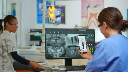 Nurse having video call with expert stomatologic medic while doctor is working with patient in background. Stomatologist assistant listening dentist using mobile webcam sitting on stomatological chair