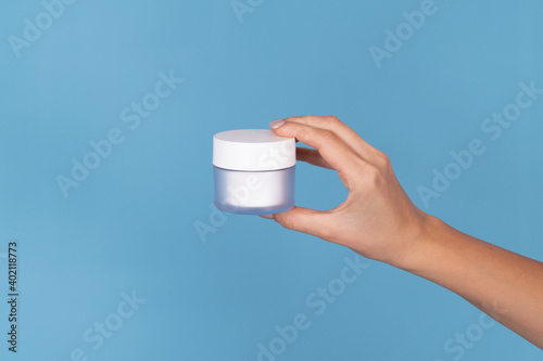 Female hand with perfect manicure holding cream bottle of lotion or face cream. Girl give jar cosmetic products on blue background with copy space