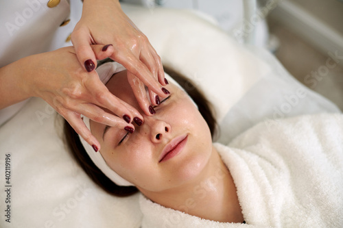 Hands of cosmetology specialist making facial lifting massage for pretty young woman in SPA salon photo