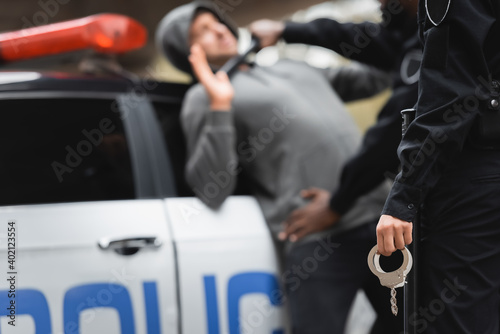 policewoman holding handcuffs with blurred multicultural policeman and offender on background outdoors.