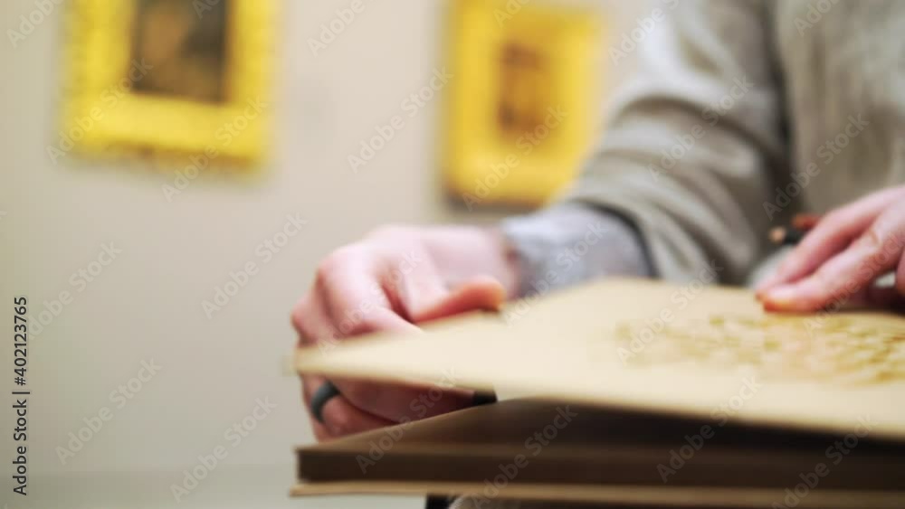 young female artist open paper notebook with brown pages. Extreme close-up artist draw in art museum at blurred background wall with artwork in golden frame young female artist open paper notebook with brown pages. Extreme close-up artist draw in art museum at blurred background wall with artwork in golden frame