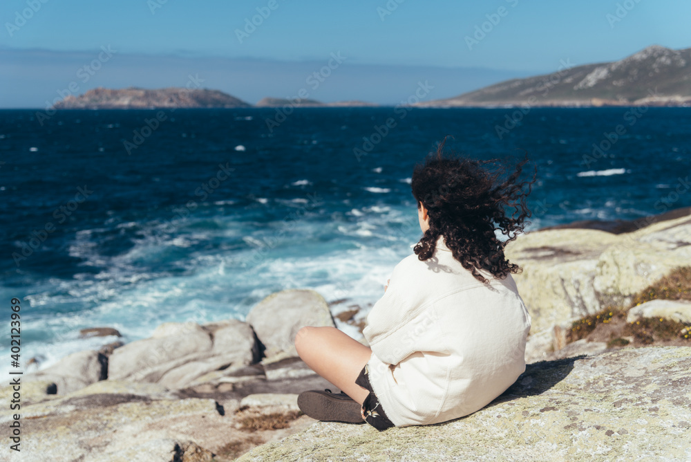 Young woman with curly hair sitting on the rocks looking at the splashing sea. View from behind