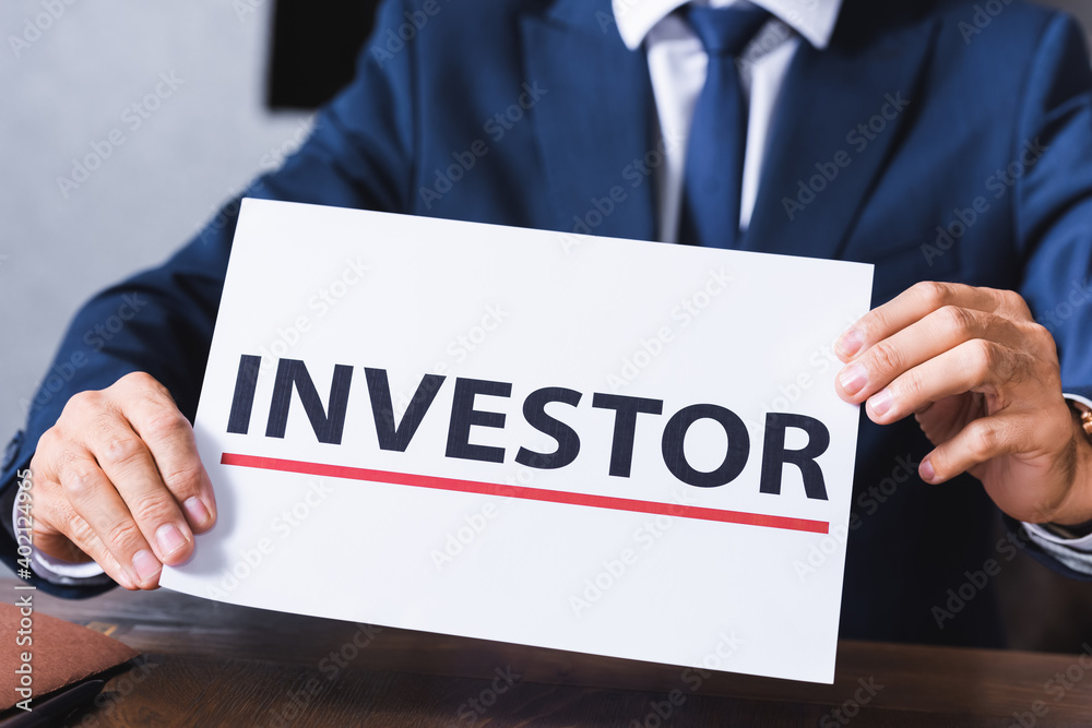 Cropped view of businessman holding sign with investor lettering in meeting room on blurred background. Cropped view of businessman holding sign with investor lettering in meeting room on blurred background.