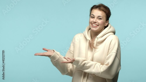 Slow motion of cute modest girl introduce product, smiling and pointing hands left at copy space, demostrate logo, standing over blue background photo