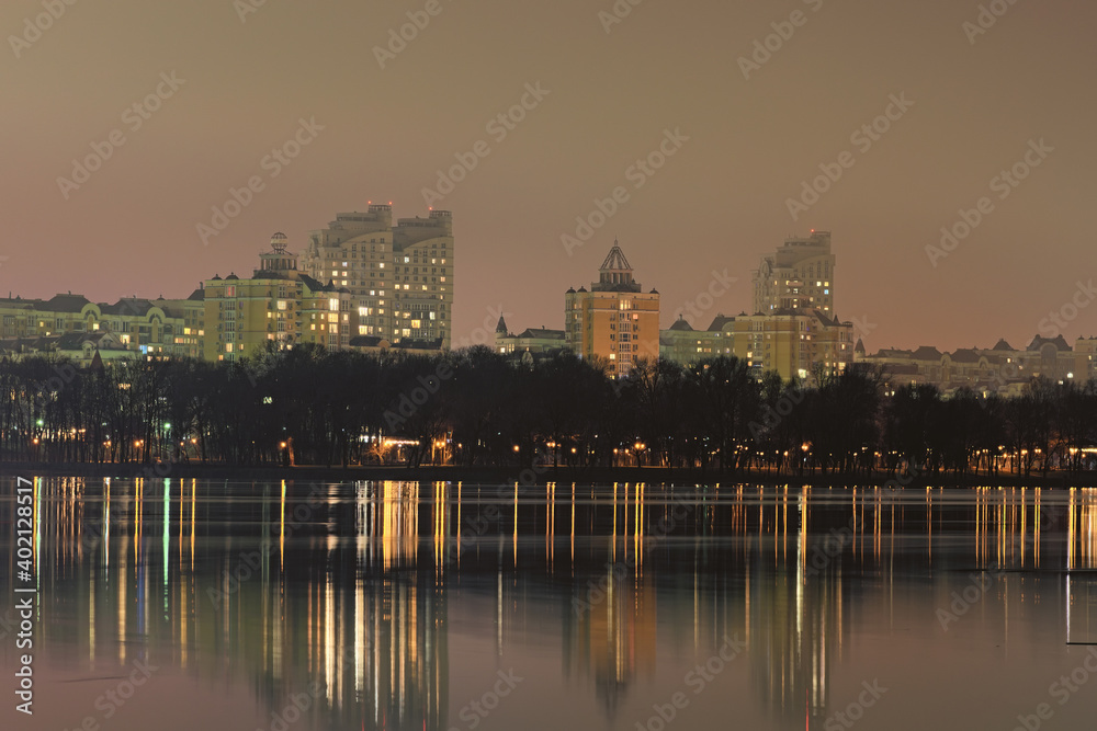 Scenic night cityscape of Obolon neighborhood with modern skyscrapers. Buildings reflected in the tranquil water of Dnieper River. Embankment with renovated Natalka Park. Kyiv, Ukraine