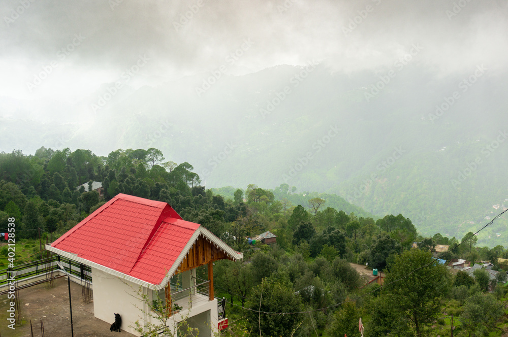 Red roofed house residence hotel homestay lodge looking over a cloudy fog filled valley with trees fading off into the distance shows the beauty of hill stations like shimla Mcleodganj 