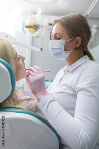 Vertical shot of female dentist in medical face mask working, examining teeth of female patient