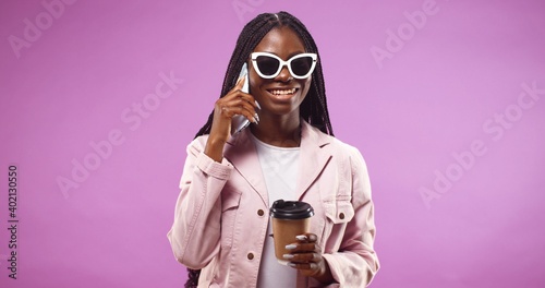 Portrait of smiling African American girl talking on mobile phone isolated over pink background. Black woman using smartphone while laughing and holding paper cup with coffee. © VAKSMANV