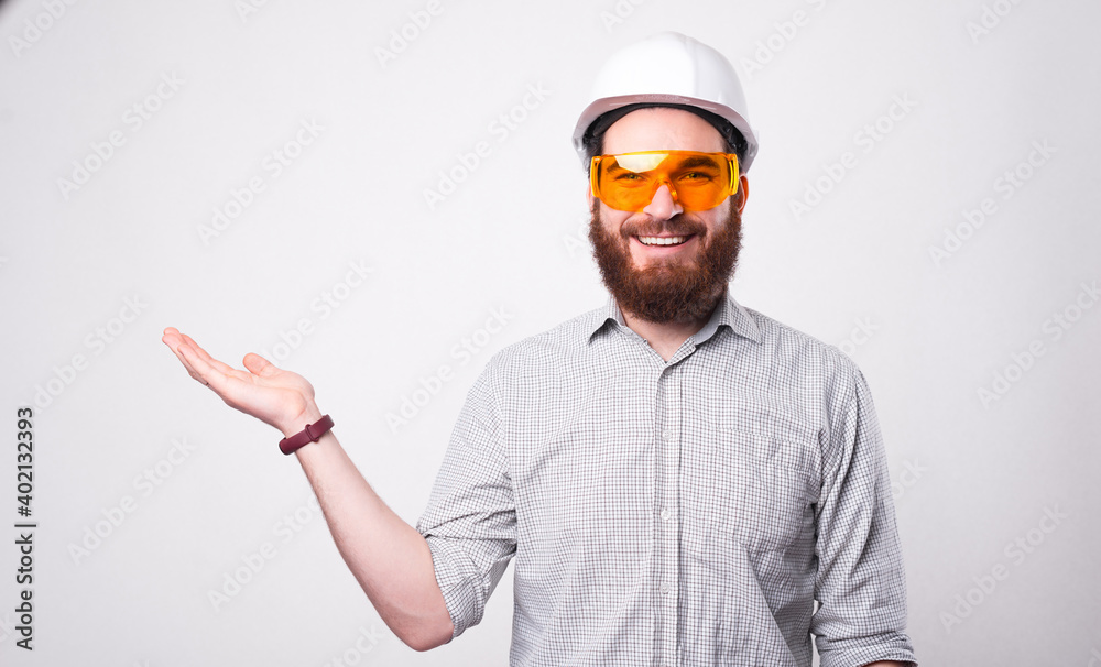Photo of cheerful handsome architect man wearing helmet and protective glasses holding copyspace in palm. Photo of cheerful handsome architect man wearing helmet and protective glasses holding copyspace in palm.