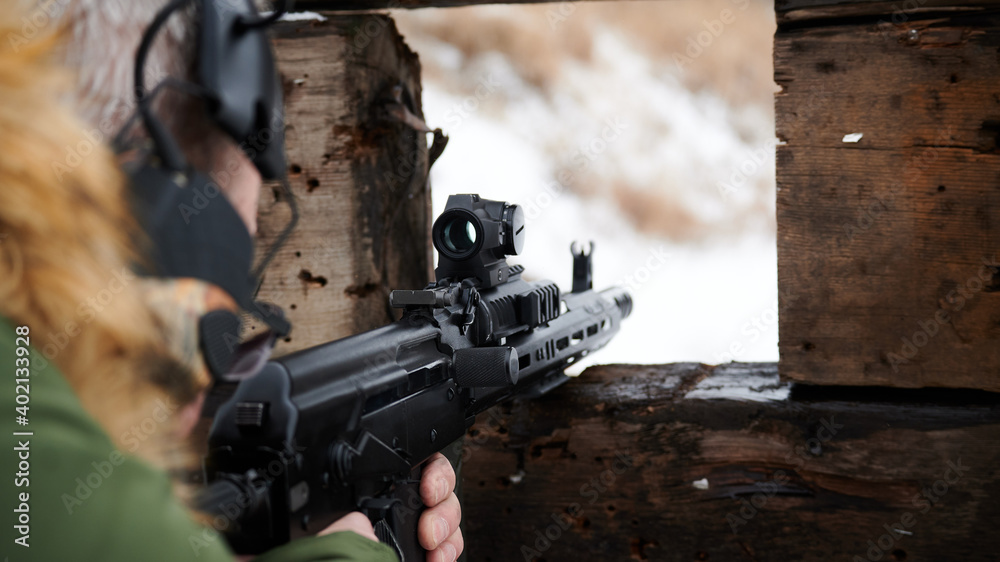Shooter aim from behind a wooden shelter through a special optical callmiator with hearing protection in a jacket in winter Shooter aim from behind a wooden shelter through a special optical callmiator with hearing protection in a jacket in winter