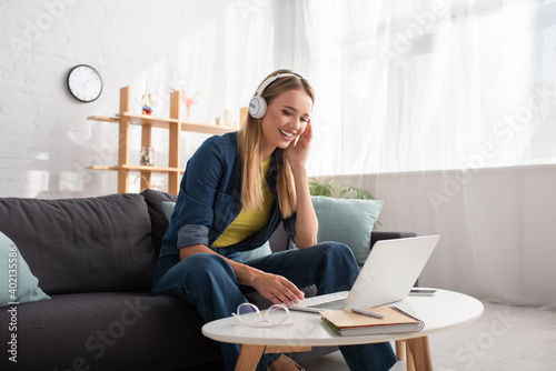  young blonde woman in headphones looking at laptop while sitting on couch at home